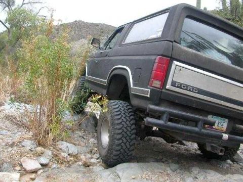1984 Ford Bronco - This black Ford Bronco, likely from the early 1980s, navigates rocky terrain with its large off-road tires visible.