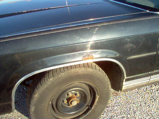 Black car fender with chrome trim and rust spot above the wheel. The tire is worn