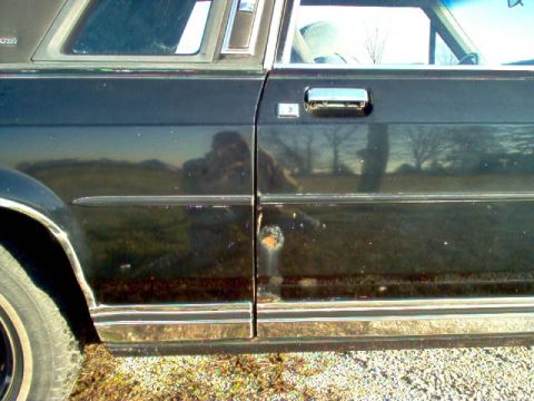 1985 Ford LTD Crown Victoria - A close-up shot of a black sedan's side door, revealing chrome accents and a small patch of rust.