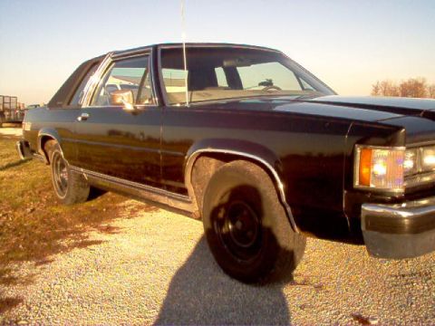 1985 Ford LTD Crown Victoria - This black sedan is seen from the front quarter angle, parked on a gravel surface under a clear sky.