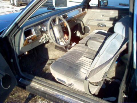 1985 Ford LTD Crown Victoria - Interior view through the open driver's door of an older sedan, showing gray velour bench seat, wood-grain dash trim, steering wheel, and w…
