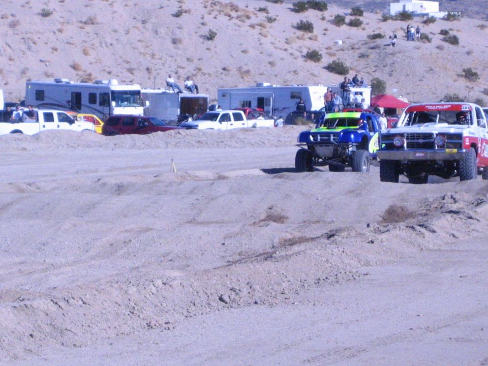 Green, blue and red off-road race trucks on a dirt track near RV pits