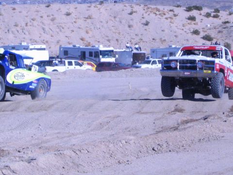 1990 Ford Bronco - blue off-road race truck and a red-and-white truck kick up dust racing across a desert dirt course.