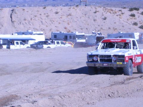 1990 Ford Bronco - red-and-white No. 308 off-road race truck drives a dirt course in front of a row of RVs in the desert.