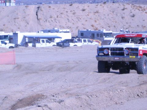 1990 Ford Bronco - red-and-white No. 308 off-road race truck with round headlights rounds a dirt track past RVs in the desert.
