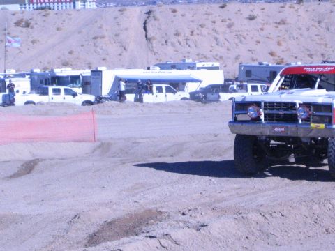 1990 Ford Bronco - front of a red-and-white off-road race truck enters a dirt course past an orange fence and RV pits in the desert.