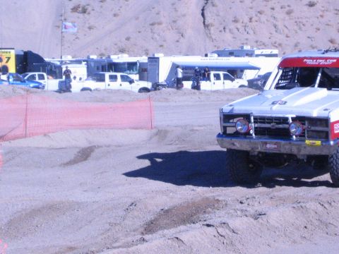 1990 Ford Bronco - red-and-white off-road race truck with twin round headlights rounds a dirt track past RVs and an orange fence.
