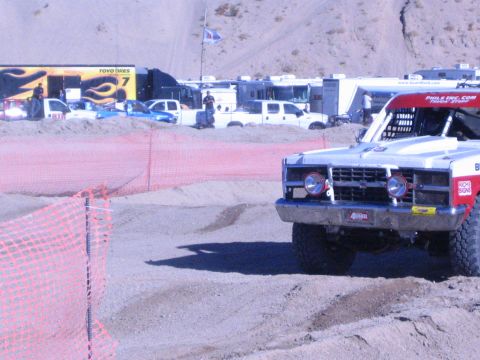 1990 Ford Bronco - red and white off-road race truck waits on a dirt track lined with orange fencing at a desert race venue.