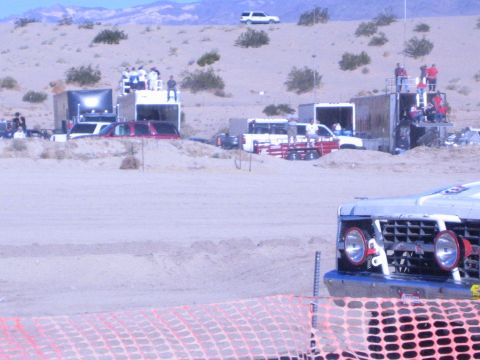 1990 Ford Bronco - Spectators stand on platforms among trailers in the desert pits while a race truck sits behind orange fencing.