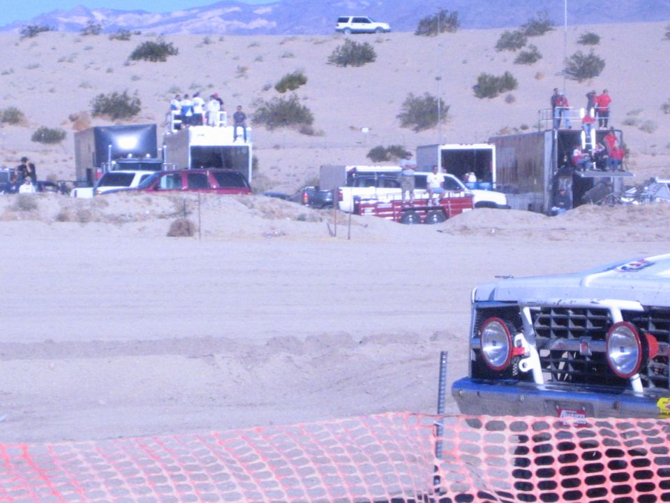 Desert race pits with spectators on platforms and a race truck in foreground