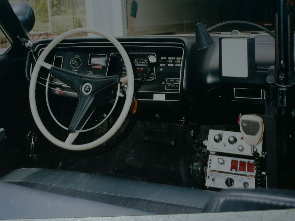 Interior dash of an older vehicle with white three-spoke steering wheel and mounted radio equipment