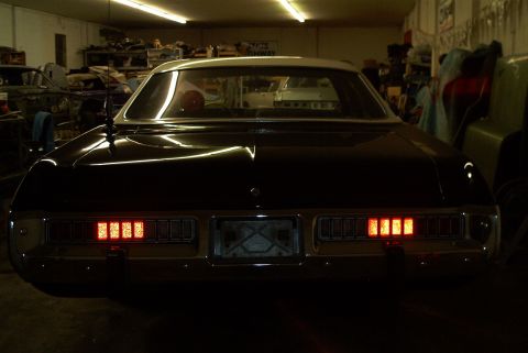 1991 Ford LTD Crown Victoria - Rear of a dark full-size sedan with its full-width taillights illuminated, parked inside a cluttered workshop.