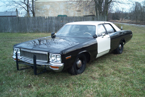 Front three-quarter view of a black-and-white police-style sedan parked on grass by a fence