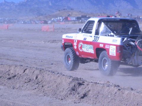 1990 Ford Bronco - Red and white off-road race truck number 301 powers along a rutted dirt course with desert mountains behind.