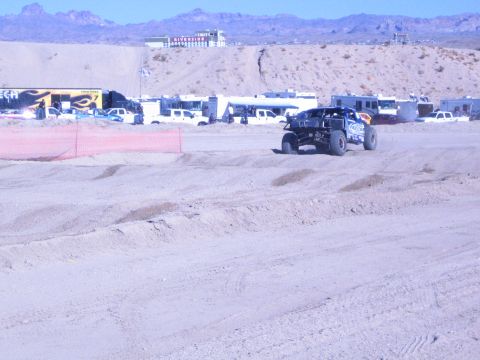1990 Ford Bronco - dark off-road race truck crests a sandy berm on a desert course with trailers and a hilltop building behind.