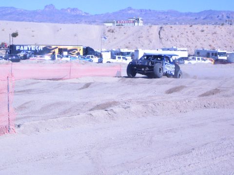 1990 Ford Bronco - white off-road race truck rounds a sandy desert course past a FABTECH banner with mountains in the distance.