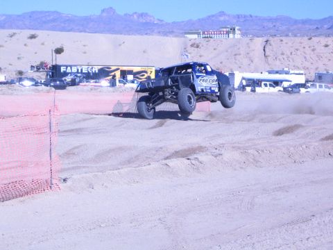 1990 Ford Bronco - white and blue off-road race truck goes airborne over a berm on a desert course with a FABTECH banner behind.
