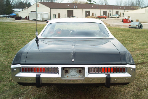 Rear view of a dark full-size sedan with full-width taillights parked outside near buildings
