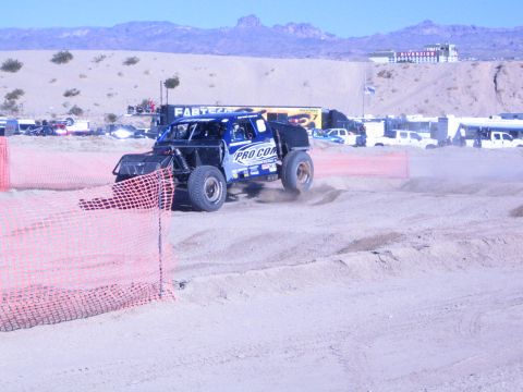 1990 Ford Bronco - blue Pro Comp off-road race truck races along a sandy desert course past trailers and a hilltop building.