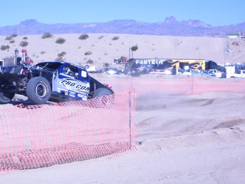 1990 Ford Bronco - blue Pro Comp off-road race truck rounds a sandy track behind orange fencing with a FABTECH banner in the back.