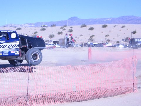 1990 Ford Bronco - blue Pro Comp off-road race truck throws up dust on a sandy desert course lined with orange fencing.