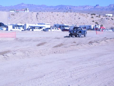 1990 Ford Bronco - dark off-road race truck drives a wide sandy course with rows of trailers and RVs in the desert pits behind.