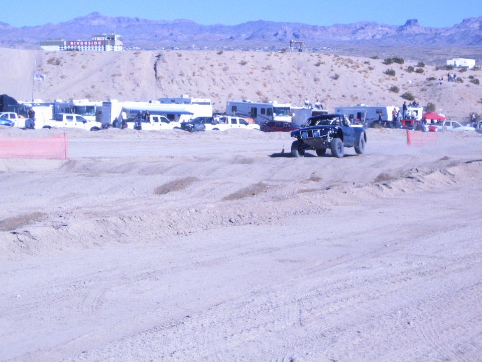 Dark off-road race truck on a wide dirt course with desert pits behind