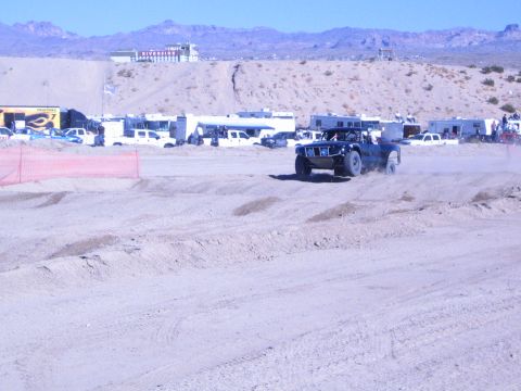 1990 Ford Bronco - dark off-road race truck raises a dust trail on a sandy desert course with trailers and mountains beyond.