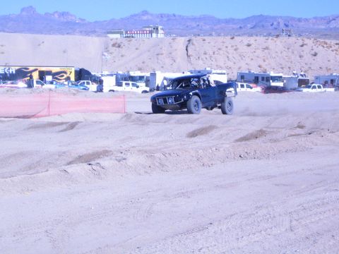 1990 Ford Bronco - dark blue off-road race truck rides a rutted sandy course with desert hills and a hilltop building behind.