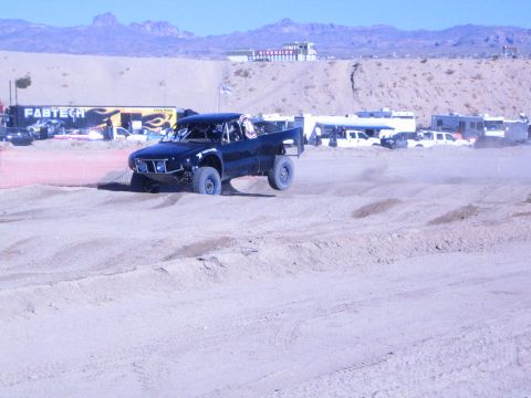 1990 Ford Bronco - dark blue off-road race truck climbs a sandy berm on a desert course past a FABTECH banner and pits.