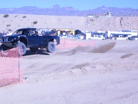 1990 Ford Bronco - dark off-road race truck kicks up dust along a sandy course lined with orange fencing in the desert.