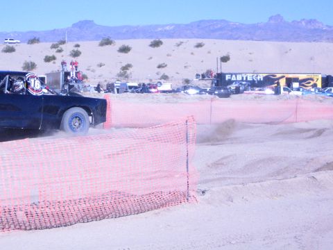 1990 Ford Bronco - dark off-road race truck moves behind orange course fencing with pits and a FABTECH banner in the background.
