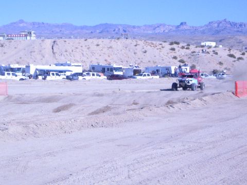 1990 Ford Bronco - red off-road race truck raises dust on a wide sandy course backed by rows of trailers and desert mountains.
