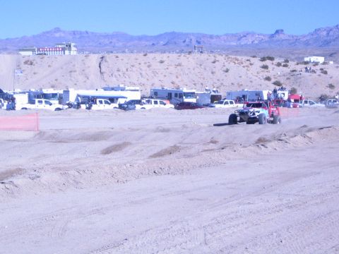 1990 Ford Bronco - off-road race buggy runs along a graded sandy desert course lined with RVs, with arid mountains on the horizon.