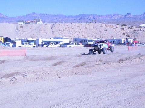 1990 Ford Bronco - off-road race buggy climbs a sandy whoop on the desert course, with pit vehicles and mountains in the distance.