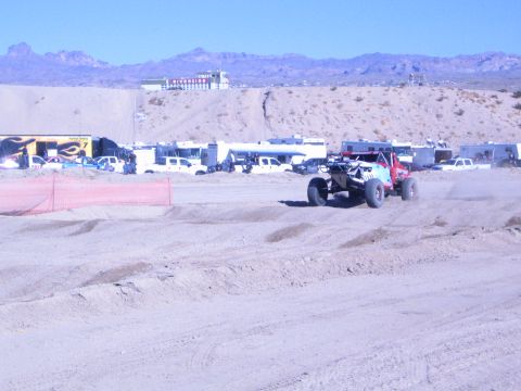 1990 Ford Bronco - red and white off-road race buggy runs the sandy desert track past orange safety fencing and parked support vehicles.
