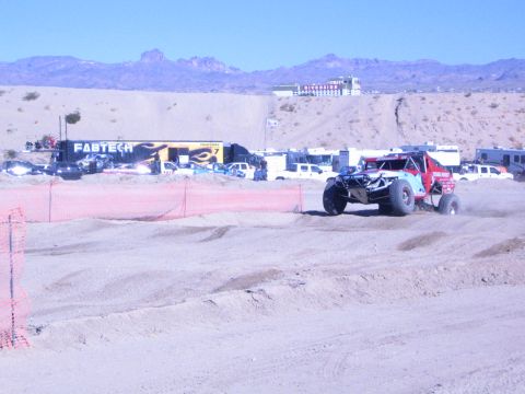 1990 Ford Bronco - red off-road race buggy runs a sandy desert course behind orange fencing, with a Fabtech display banner in the pits.