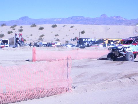 1990 Ford Bronco - off-road race buggy sits near the staging area behind orange safety fencing, with a Fabtech banner and pits behind.