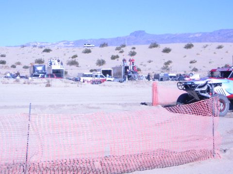 1990 Ford Bronco - race buggy is parked beside orange safety fencing in a dusty desert pit area with support trucks and trailers.