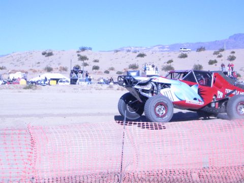 1990 Ford Bronco - red, white and teal off-road race buggy with a tall roll cage sits on the sandy desert course behind orange fencing.