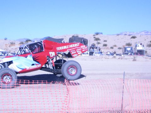 1990 Ford Bronco - red off-road race buggy marked Terrible Herbst sits on the sandy desert track with spectators and pits behind.