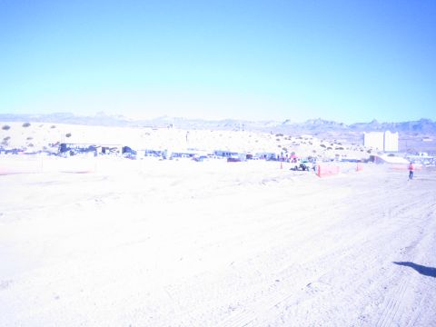 1990 Ford Bronco - bright, washed-out wide view of a desert off-road racing venue with distant pits, fencing and mountains.