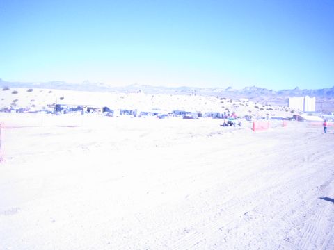 1990 Ford Bronco - washed-out wide shot of a sandy desert racing course with distant pit structures, fencing and mountains.