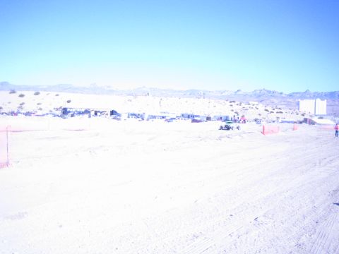 1990 Ford Bronco - bright wide view of a desert off-road race venue with a row of parked vehicles, orange fencing and mountains.