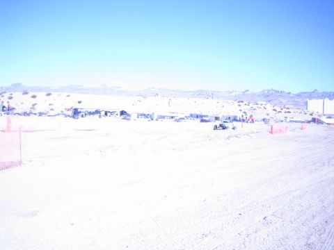 1990 Ford Bronco - washed-out wide shot of a sandy desert race course with distant pit vehicles, orange fencing and hills.