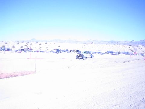 1990 Ford Bronco - small off-road race buggy runs a bright sandy desert course with orange fencing and distant pits behind.