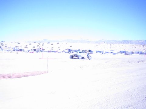 1990 Ford Bronco - off-road race buggy crosses a sandy desert course with orange fencing and a row of distant pit vehicles.