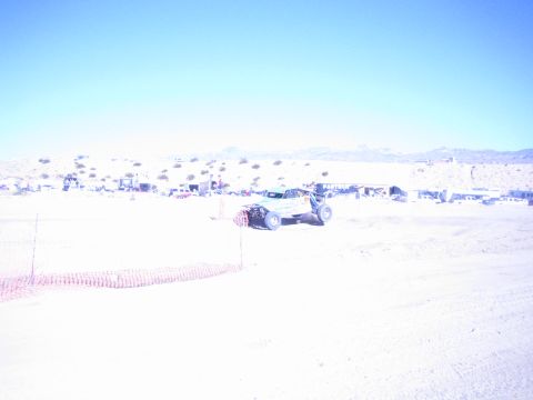 1990 Ford Bronco - light-colored off-road race buggy runs a sandy desert course behind orange safety fencing with distant pits.