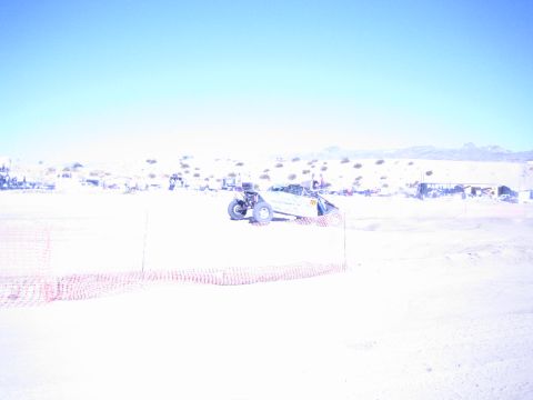 1990 Ford Bronco - numbered off-road race buggy runs a sandy desert course behind orange fencing with distant pit structures.