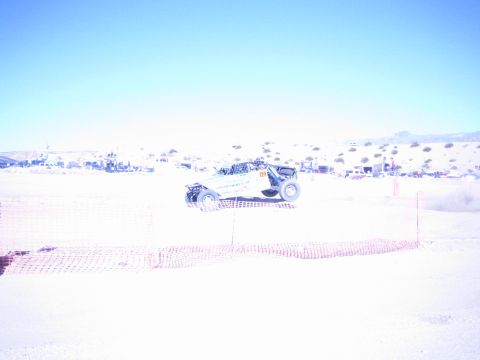 1990 Ford Bronco - white off-road race buggy kicks up dust on a sandy desert course behind orange fencing with distant pits.